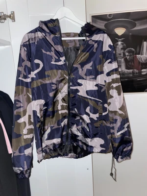 Camouflage vindjacka från Lindfors - Snygg vindjacka med camomönster i blått, grönt och grått från Lindfors Stockholm. Jackan har huva, dragkedja framtill och justerbar dragsko nedtill. Perfekt för dig som vill sticka ut med streetstyle-vibe.