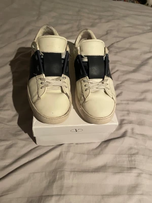 Valentino open - Snygga vita sneakers Valentino Garavani med bred blå detalj på skorn. Skorna är i ett slitet skick på flera ställen som man kan se på bilderna, men annars är dem bra om man vill sticka ut lite gran.               Diskutera pris