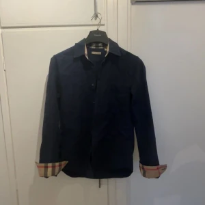 Mörkblå skjorta från Burberry Brit - Snygg mörkblå skjorta från Burberry Brit med klassiskt rutigt mönster på insidan av kragen och ärmsluten. Skjortan har knappar framtill, bröstficka och är långärmad. Perfekt för dig som gillar stilrena detaljer och ikonisk design.