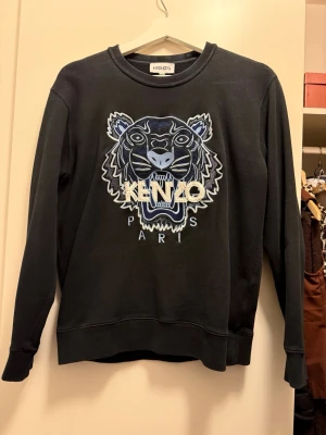 Svart Kenzo sweatshirt med tiger - Svart sweatshirt från Kenzo i 100% bomull med ikoniskt broderat tigerhuvud och Kenzo Paris-logga i blå och vita toner på bröstet. Tröjan har rund halsringning och långa ärmar, perfekt för dig som vill sticka ut med en streetig vibe.