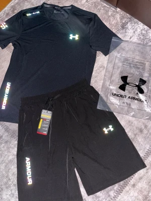 Under Armour reflactive set Size L - Bilderna visar ett svart träningsset från Under Armour med t-shirt och shorts, båda med reflekterande logga och text. Inga skor syns på bilderna.