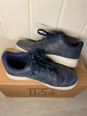 Nike Air Force 1  - Säljer ett par klassiska Nike Air Force 1 sneakers i mörkblått skinn med matchande blå snörning och vit platt sula. Modellen har perforerad tå för extra ventilation och en rund tåform. Perfekt för dig som gillar streetstyle och vill ha en ikonisk sneaker i garderoben.