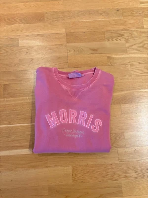 Rosa sweatshirt från Morris - Rosa sweatshirt från Morris med broderad logga och texten 'Great Britain Heritage' på bröstet. Tröjan har rund hals, ribbade muddar och lång ärm. Perfekt för dig som gillar klassisk college-stil och vill ha en färgklick i garderoben.
