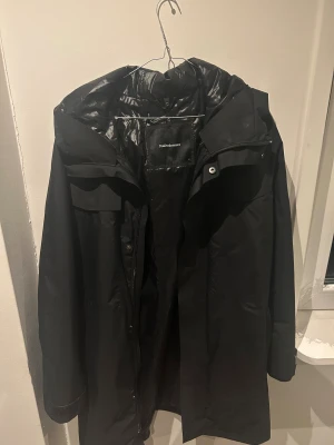Svart lång kappa av Peak Performance - Säljer en svart lång dunjacka från Peak Performance med huva och diskreta fickor. Jackan har dragkedja och knappar framtill, samt ett stilrent och minimalistiskt utseende. Perfekt för kalla dagar och har en snygg, enkel design som funkar till allt. Nypris 6400kr