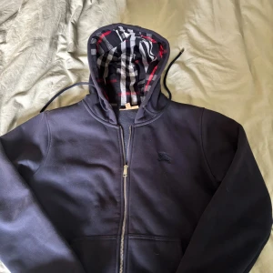 Mörkblå Burberry hoodie med dragkedja - Mörkblå hoodie från Burberry med klassiskt broderat logomärke på bröstet. Insidan av huvan har det ikoniska rutiga Burberry-mönstret i svart, vitt och rött. Tröjan har dragkedja framtill och snörning vid huvan. Perfekt för dig som vill ha en stilren och lyxig look.