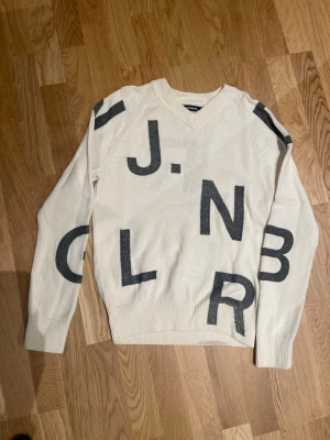 Vit stickad v-ringad tröja J.Lindeberg S män - Vit stickad tröja från J.Lindeberg i storlek S. Tröjan har ett coolt print med stora svarta bokstäver över hela plagget och v-ringad hals. Långa ärmar och ribbade muddar vid ärmslut och nederkant. Perfekt för dig som gillar statement-plagg.