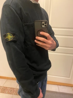 Svart Stone Island sweatshirt XL - Svart sweatshirt från Stone Island i storlek XL. Klassisk design med rund hals, bröstficka och ikonisk patch med logga på vänster ärm. Tröjan har långa ärmar och ribbade muddar. Perfekt för dig som gillar streetwear och vill ha en clean look.