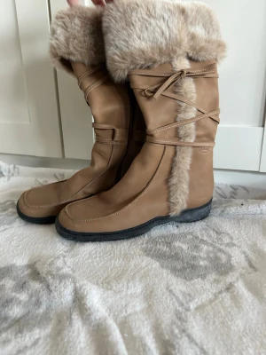 Beiga Scholl boots med pälsdetalj - Säljer ett par snygga beiga boots från Scholl med mjuk päls runt skaftet och längs sidan. Skorna har rund tå, platt sula och dekorativa snören som går runt skaftet. Perfekta för kalla dagar och har en mysig vinterlook.