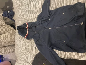 Mörkblå vindjacka från Moncler - Snygg mörkblå vindjacka från Moncler med huva och coola rödvita ränder på kanten. Jackan har dragkedja framtill, två fickor med knappar och Moncler-logga på ärmen. Perfekt för dig som vill ha en stilren och sportig look.