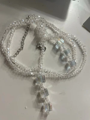 Långt halsband ny  - Bilden visar ett halsband med transparenta pärlor och kubformade detaljer, 