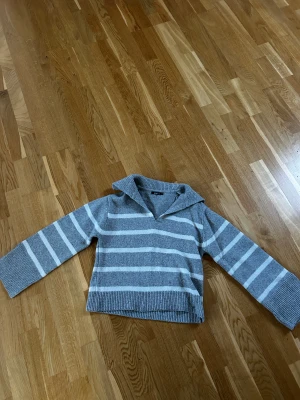 Grå stickad tröja med vita ränder  - Grå stickad tröja med vita ränder och krage i ett mjukt och skönt material. Slitsar på armarna ger en snygg detalj. Inga skavanker.
