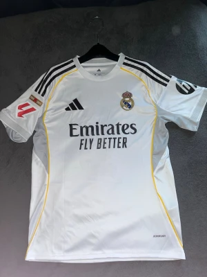 Real Madrid Mbappé matchtröja Adidas - Säljer en vit Real Madrid matchtröja från Adidas med Mbappé och nummer 9 på ryggen. Tröjan har svarta detaljer, gula linjer längs sidorna och klubbmärke på bröstet. Tillverkad i lätt polyester med AEROREADY-teknologi och klassiska Adidas-ränder på axlarna. Köpte tröjan tidigt varav felprintet.