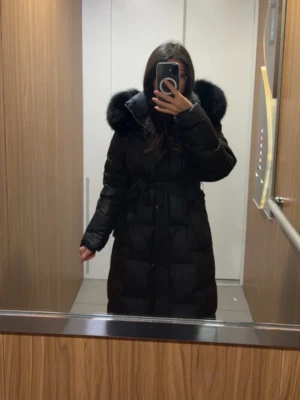 Svart jacka  - Lång svart pufferjacka med stor huva och bred päls runt kanten. Jackan har bälte i midjan för snygg passform och stängs med dragkedja och knappar. Perfekt för kalla dagar och riktigt mysig att bära.