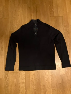 Mörkblå stickad tröja från Ziprin - Snygg mörkblå stickad tröja från Ziprin med ribbad krage och knappdetaljer vid halsen. Tröjan har en liten bröstficka och långa ärmar, perfekt för dig som gillar stilren och enkel design. Passar dig som vill ha en chill och klassisk look.