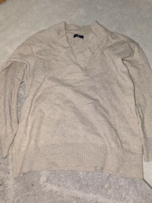 Beige stickad v-ringad tröja GANT - Snygg beige stickad tröja från GANT med v-ringad hals och långa ärmar. Materialet har en lätt glittrig finish som ger en extra touch. Perfekt för dig som gillar klassisk stil med lite edge. Ribbstickade muddar vid ärmslut och nederkant.