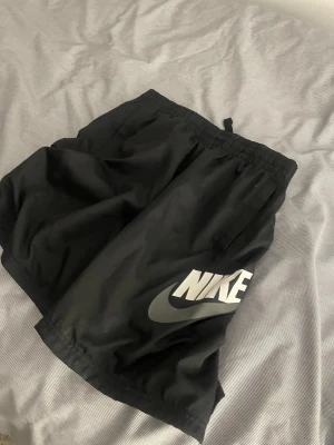 Svarta Nike shorts XL - Svarta shorts från Nike i storlek XL med stor vit Nike-logga på ena benet. De har elastisk midja och är tillverkade i ett lätt och luftigt syntetmaterial, perfekt för träning eller chill. Klassisk sportig look med fickor på sidorna.pris kan diskuteras genom snabb affär