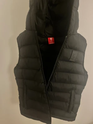 Svart dunväst från Liverpool FC - Svart dunväst från Liverpool FC med huva och diskret klubbmärke på bröstet. Västen är quiltad och har dragkedja framtill samt två fickor. Perfekt för kyliga dagar när du vill visa vilket lag du hejar på. Oanvänd, utan prislapp.