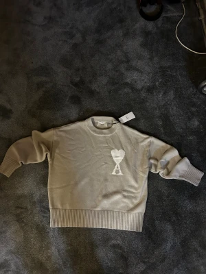 Beige stickad tröja från Ami Paris - Snygg beige stickad tröja från Ami Paris med det ikoniska hjärta-A broderiet på bröstet. Tröjan har rund hals, ribbade muddar och nederkant samt långärmad passform. Perfekt för dig som gillar stilrena och trendiga plagg.
