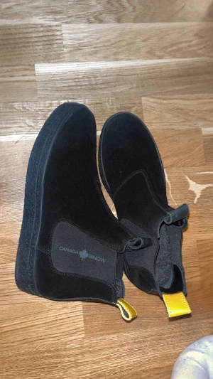 Svarta Chelsea boots från Canada Snow - Svarta Chelsea boots från Canada Snow med mjuk mocka på utsidan och värmande ullblandning på insidan. Snygga elastiska paneler på sidorna och gul dragflik bak. Perfekt för kalla dagar med platt sula och rund tå.