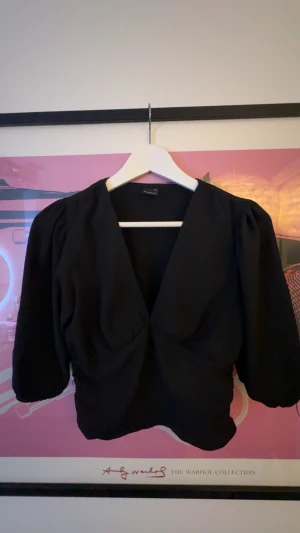 Svart omlottblus med puffärm - Svart blus från Gina med djup v-ringning. Toppen har trekvarts puffärmar och smockad midja som ger en snygg siluett. Materialet är lätt strukturerat och känns mjukt mot huden. Perfekt för dig som gillar stilrena och trendiga plagg. Liten fläck men kan nästan garantera att den går bort i tvätten.