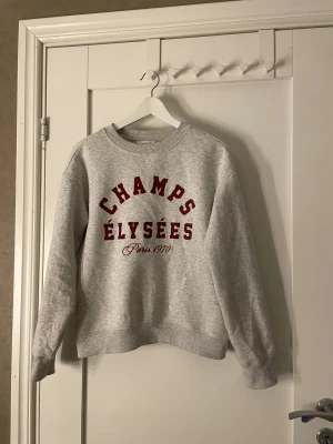 Grå sweatshirt Champs Élysées H&M XS - Grå sweatshirt från H&M i storlek XS med röd text 'CHAMPS ÉLYSÉES Paris 1970' framtill. Tröjan har rund hals, och är långärmad. Perfekt för dig som gillar enkel och snygg streetstil.