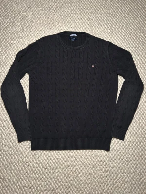Svart kabelstickad tröja från GANT - Svart kabelstickad tröja från GANT i storlek L men passar även Medium. Tröjan har rund hals, ribbade muddar och ett broderat GANT-logo på bröstet. Materialet är mjuk bomull och den har långärmad passform. Perfekt för dig som gillar klassisk och stilren look.