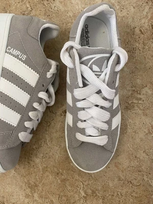 Grå Adidas Campus sneakers - Snygga Adidas Campus sneakers i ljusgrå mocka med klassiska vita ränder och vit sula. Skorna har rund tå, platt sula och vita snören. Perfekt för dig som gillar stilrena och tidlösa sneakers med retrovibe.