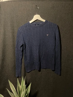 Marinblå kabelstickad tröja från GANT - Snygg marinblå kabelstickad tröja från GANT med rund hals och diskret logga broderad på bröstet. Tröjan har långa ärmar och ribbade muddar vid hals, ärmslut och nederkant. Perfekt för dig som gillar klassisk och stilren look.