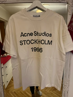 Vit Acne Studios t-shirt Stockholm 1996 - Vit t-shirt från Acne Studios med svart tryck 'Acne Studios STOCKHOLM 1996' på framsidan. Klassisk passform, rund hals och korta ärmar. Tillverkad i 100% bomull för en skön och avslappnad vibe.