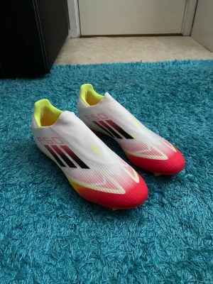 Adidas F50 fotbollsskor röd/vit - Adidas F50 fotbollsskor i en riktigt snygg färgkombo med vit ovandel, röda och gula detaljer samt svarta ränder. Skorna har platt sula med dobbar för gräsplan och är tillverkade i syntetmaterial. Perfekt för dig som vill sticka ut på planen.