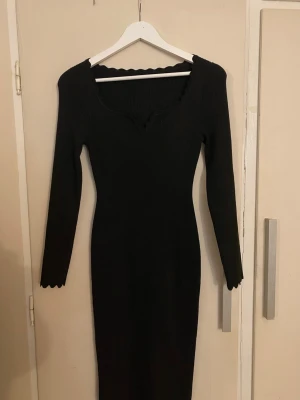 Svart ribbad långklänning med vågig kant - Svart långärmad ribbad klänning med figurnära passform. Klänningen har en snygg v-ringning och vågiga kanter vid halsringning, ärmslut och nederkant. Perfekt för dig som gillar stilrena och enkla plagg med en twist.