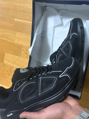 Svarta diors storlek 42 - Säljer ett par svarta Dior sneakers med silvriga detaljer och tydlig CD-logga på sidan. Skorna har snörning, platt sula och rund tå. Materialet är en mix av mesh och syntet, vilket ger dem en sportig och modern vibe.   