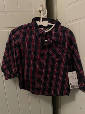 Rutig skjorta från MyWear Baby - Snygg rutig skjorta i mörkblått och rött från MyWear Baby. Skjortan har klassisk krage, knäppning framtill och en bröstficka. Perfekt för dig som gillar en stilren men ändå cool look.