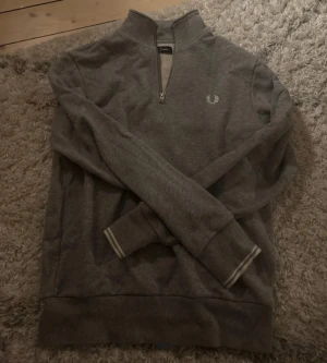 Grå half zip tröja från Fred Perry - Snygg grå långärmad tröja från Fred Perry med half zip och broderad logga på bröstet. Tröjan har ribbade muddar med vita detaljer och är gjord i mjuk bomull. Perfekt för dig som gillar stilrena och klassiska plagg. Knappt använd 