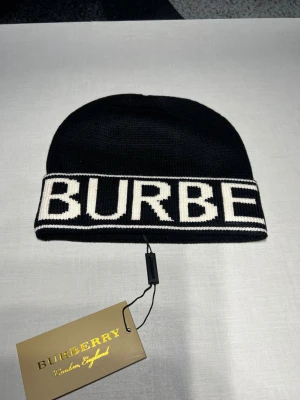 Svart Burberry mössa med logotyp - Snygg svart mössa från Burberry med bred vit logotyp runt hela kanten. Mössan är stickad i mjukt material och har en klassisk, rund form. Perfekt för dig som vill ha en stilren accessoar med tydlig designer-touch.