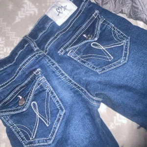 Blå jeans med vit kontrastsöm - Snygga blå jeans från Rock & Republic med vit kontrastsöm och dekorativa bakfickor med lock och knapp. Klassisk femficksmodell med coola detaljer som sticker ut. Perfekt för dig som gillar statement-jeans.