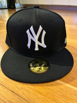 Svart New Era 59FIFTY keps - Snygg svart New Era 59FIFTY keps med klassiskt New York Yankees broderat vitt logotyp framtill. Kepsen har en rak skärm och MLB-logga baktill. Tillverkad i slitstark bomull, perfekt för dig som vill ha en clean och sportig look.