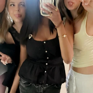 Svart blus med knappar och volang - Säljer en svart blus med dekorativa vita knappar framtill och volang nedtill. Toppen har smala axelband med rosett-detalj och en feminin, något croppad passform. Perfekt för dig som gillar en söt och trendig stil.