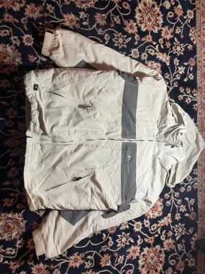Champion jacka  för Champs 💪🔥 - Snygg vindjacka från Champion i beige och grått med huva och dragkedja framtill. Jackan har två snedställda fickor med dragkedja och coola grå paneler på bröst och ärmar. Materialet är nylon och polyester, perfekt för blåsiga dagar.