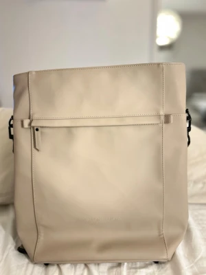 Beige ryggsäck från Gaston Luga - Snygg beige ryggsäck från Gaston Luga med stilren design och praktiska fack. Väskan har breda, justerbara axelremmar och en stor dragkedjeficka framtill. Materialet är slätt och känns lyxigt, perfekt för dig som gillar minimalistisk stil.