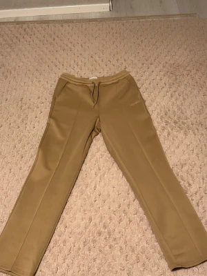 Beige mjukisbyxor från Les Deux - Säljer ett par snygga beige mjukisbyxor från Les Deux med raka ben och snörning i midjan. Byxorna har diskret logga på ena benet och är tillverkade i ett mjukt material som känns skönt mot huden. Perfekta för en chill och stilren look. HELT NYA, MED KVITTOT. original priset,1100kr