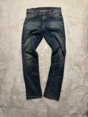 Nudie jeans slim fit - Säljer ett par Nudie jeans i blå denim med snygg slitning och smal passform. Perfekta för dig som gillar en modern och stilren look.