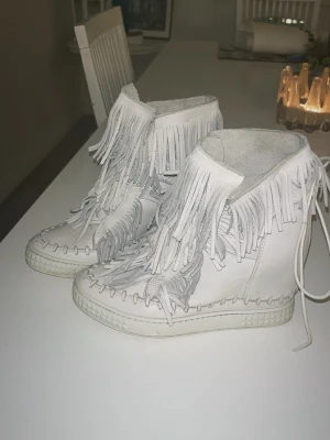 Vita mockasneakers med fransar - Unika vita sneakers i mocka med högt skaft och coola fransdetaljer runt hela skon. Skorna har snörning och en platt sula, vilket ger en avslappnad men ändå trendig look. Perfekta för dig som vill sticka ut med din stil. Dem är använda typ 2 gånger så dem är väldigt fräscha och ser nya ut. 