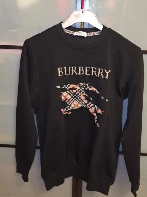 Svart Burberry sweatshirt  - Svart sweatshirt från Burberry med klassisk logga och ett stort motiv i Burberry-rutigt mönster på bröstet. Tröjan har rund halsringning, långa ärmar och ribbade muddar vid ärmslut och nederkant. Perfekt för en trendig och avslappnad stil. Den är oanvänd men den är tvättad. Tveka inte för att ställa frågor! Priset är inte hugget i sten.