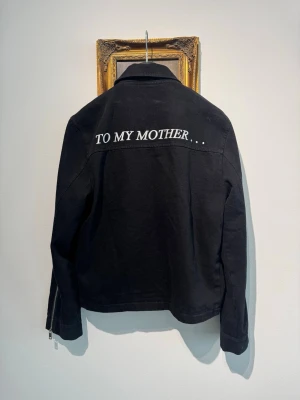 Svart jeansjacka från Enfants Riches Déprimés - Svart jeansjacka med rak passform och dragkedja framtill från Enfants Riches Déprimés. Jackan har texten 'TO MY MOTHER...' broderad i vitt på ryggen och två sidofickor. Klassisk krage och långa ärmar ger en clean och stilren vibe.