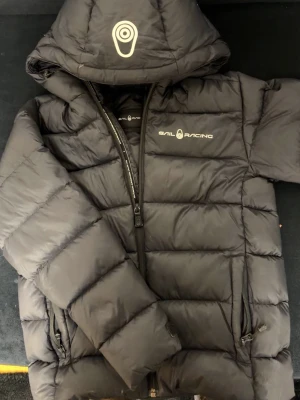 Svart dunjacka från Sail Racing - Snygg svart dunjacka från Sail Racing med huva och logga på bröstet och huvan. Jackan har quiltad design, dragkedja framtill och två sidofickor med dragkedja. Perfekt för kalla dagar och riktigt stilren look.