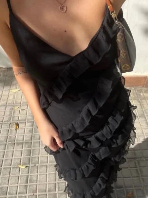 Svart volangklänning med smala band - Säljer en svart långklänning med smala axelband och v-ringning. Klänningen har flera diagonala volangdetaljer som ger en cool och trendig look. Perfekt för dig som vill sticka ut lite extra. Materialet känns lätt och flowy.