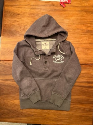 Grå hoodie från Hollister - Snygg grå hoodie från Hollister med trycket 'Hollister Surfboards' på bröstet. Hoodien har knappar vid halsen, dragsko i huvan och en stor magficka. Tillverkad i mjukt bomullsmaterial och har ribbade muddar vid ärmar och midja. Perfekt för en avslappnad stil.