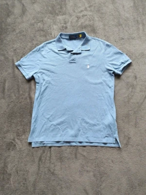 Ljusblå pikétröja Ralph Lauren - Klassisk ljusblå pikétröja från Polo Ralph Lauren med broderad logga på bröstet. Tröjan har krage, korta ärmar och knappar vid halsen. Riktigt fina färger passar perfekt till våren och sommaren 