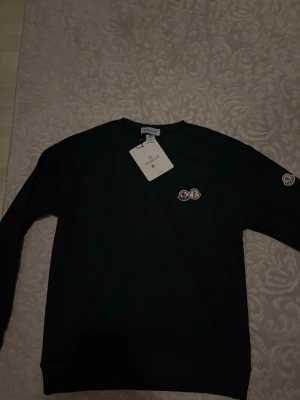 Svart sweatshirt från Moncler - Svart sweatshirt från Moncler med rund halsringning och Moncler-logga broderad på bröstet samt ärmen. Tröjan har en klassisk passform och är tillverkad i mjukt bomullsmaterial. Perfekt för en stilren och avslappnad look.